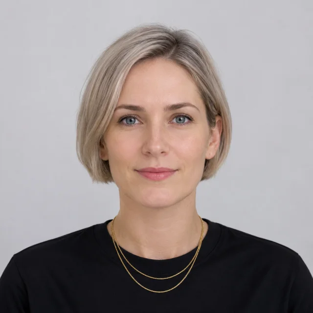Adult with a short blonde bob wears two layered gold necklaces over a black top, full face in frame.