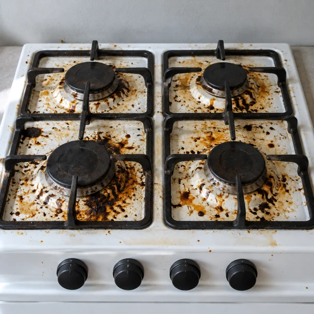 A white gas stovetop covered in grease and burnt residue, with four blank black knobs.