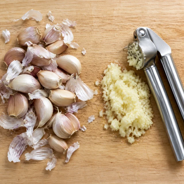 Same wood cutting board: whole garlic and skins on the left, pressed garlic and a stainless garlic press on the right.