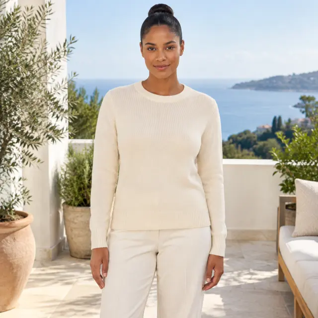 An adult woman with a tight bun wears a plain cream knit top on a bright terrace.