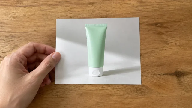 Hand holding a borderless print of a mint cleanser tube over a wood desk.