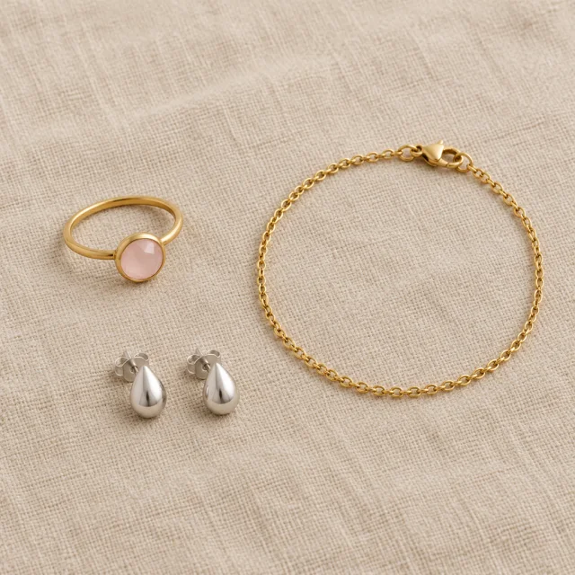 A pink-stone ring, a pair of silver teardrop studs, and a gold chain bracelet lie flat on beige linen.
