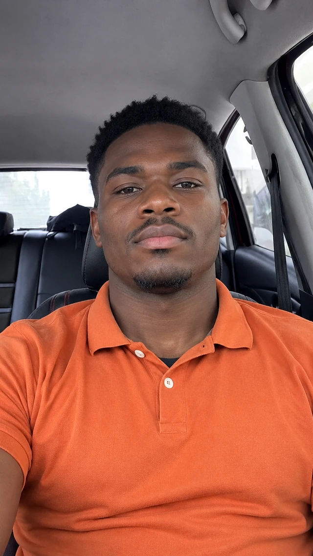 Adult Black man with short sponge curls in a car setting, speaking to camera seated in the parked passenger seat giving a casual car-selfie introduction.