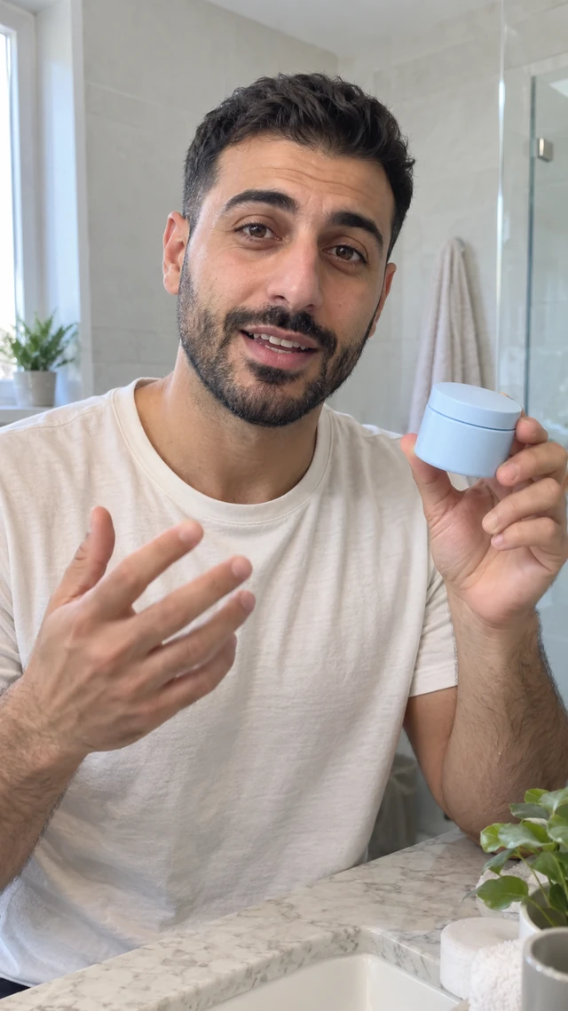 Adult Middle Eastern man with a short dark crop explains a plain blue moisturizer jar at a bright bathroom counter.