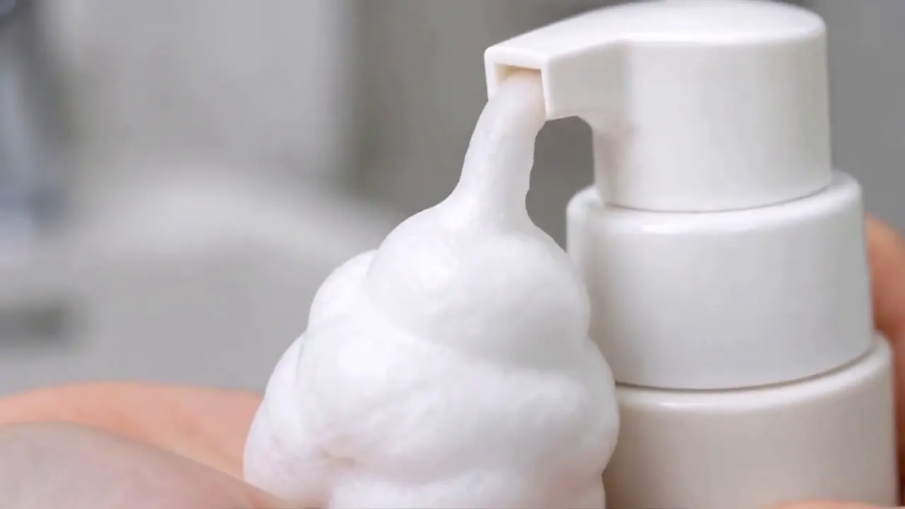 A hand presses a white foaming cleanser pump and spreads foam while washing a face.