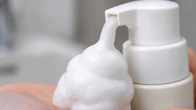 A hand presses a white foaming cleanser pump and spreads foam while washing a face.
