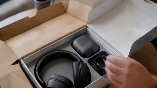 Hands open a gray headphone package and remove the headphones and charging case.