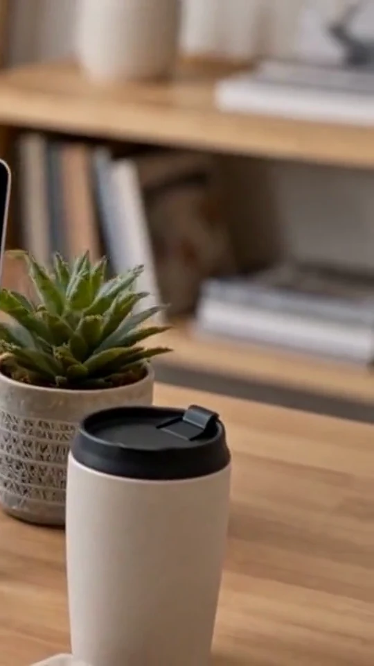 A white travel mug sits beside a plant and books while a product page is open.