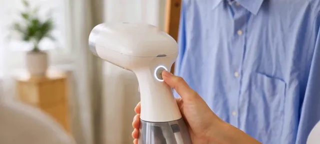 Hand pressing the power button of a white handheld garment steamer in front of a blue shirt