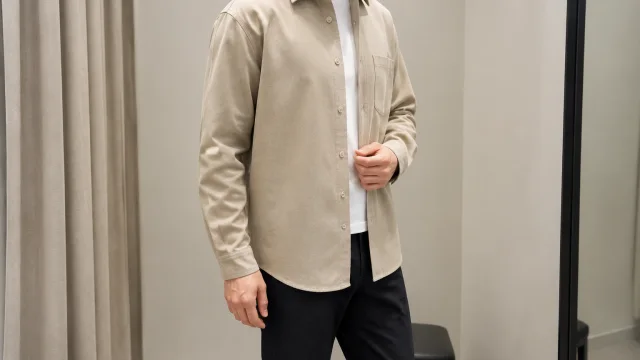 Adult wearing a plain beige overshirt and dark trousers in a bright hallway.