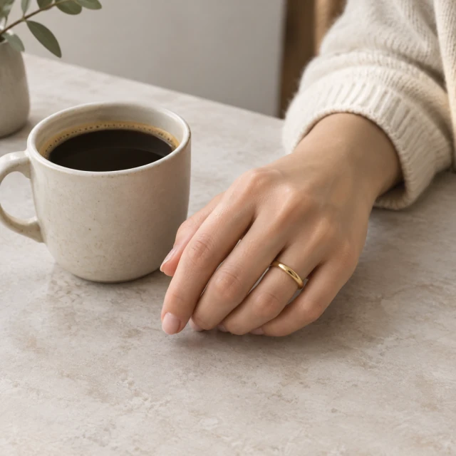Gold ring on an adult hand beside a ceramic coffee cup in soft window light.