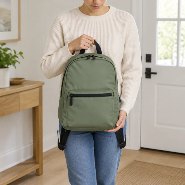 Adult shown from shoulders down holding a compact backpack at hip height in a bright entryway.