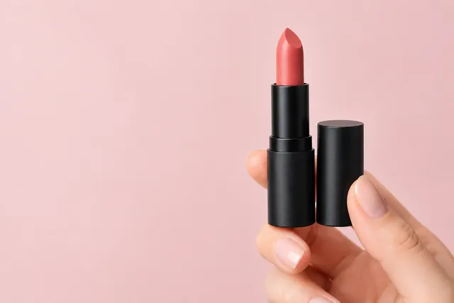 Hand holding a coral lipstick and its cap against a soft pink backdrop with open space on the left