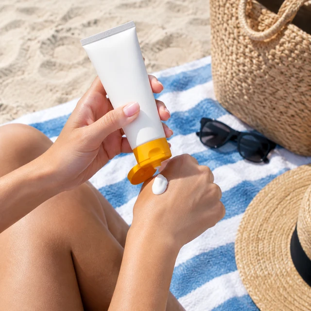 Adult holds a sunscreen tube beside a beach towel before heading out