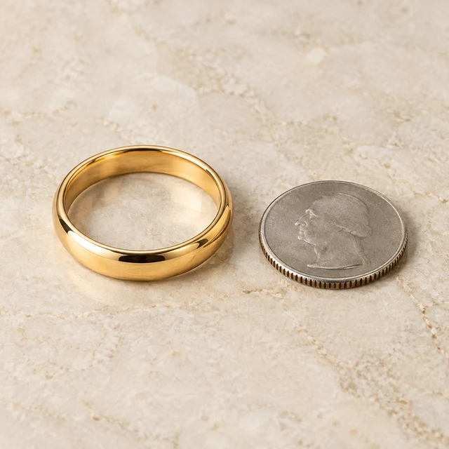 Plain polished yellow-gold band ring beside a small neutral metal coin on a warm pale stone surface.