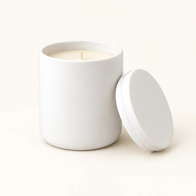 White ceramic candle jar with an open rim, visible wax, wick, and lid on a warm-white background.