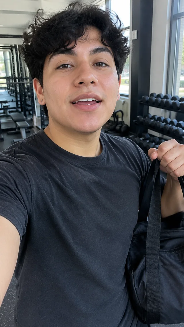 an adult Latino nonbinary person in their early twenties in a gym setting speaks to camera while holding a plain plain black gym duffel.