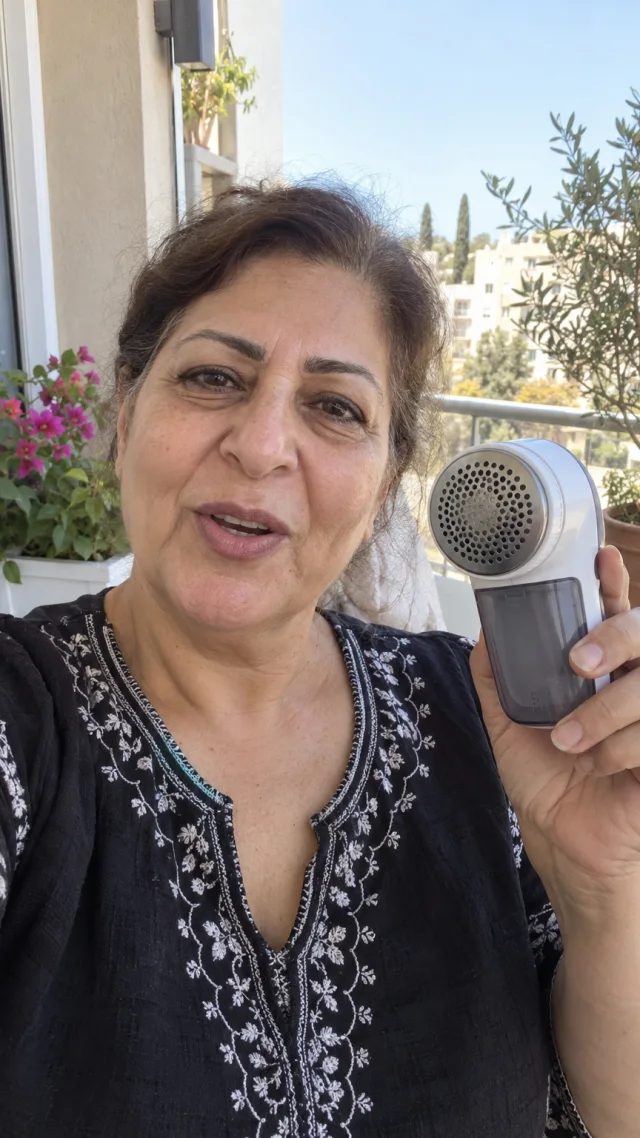 An adult Middle Eastern woman in her late fifties on a balcony speaks to camera while holding a plain handheld fabric lint remover.