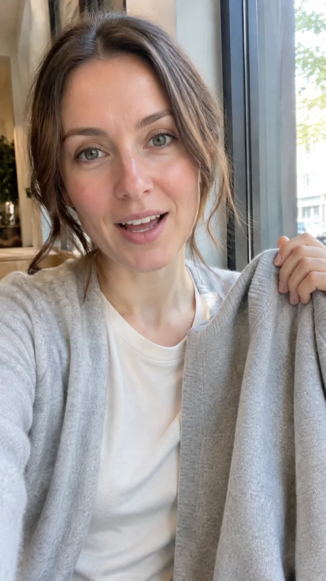 an adult white woman around thirty in a cafe setting speaks to camera while holding a plain soft everyday cardigan.