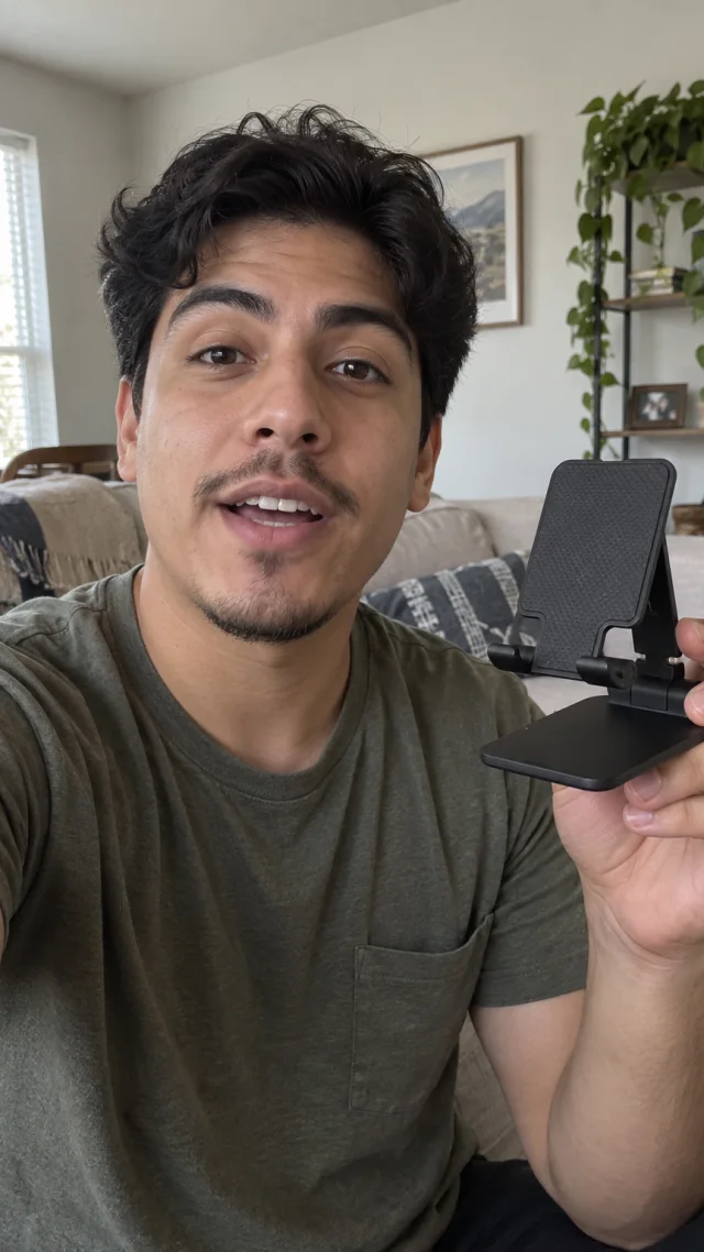 an adult Latino man in his early twenties in a living room setting speaks to camera while holding a plain folding smartphone stand.