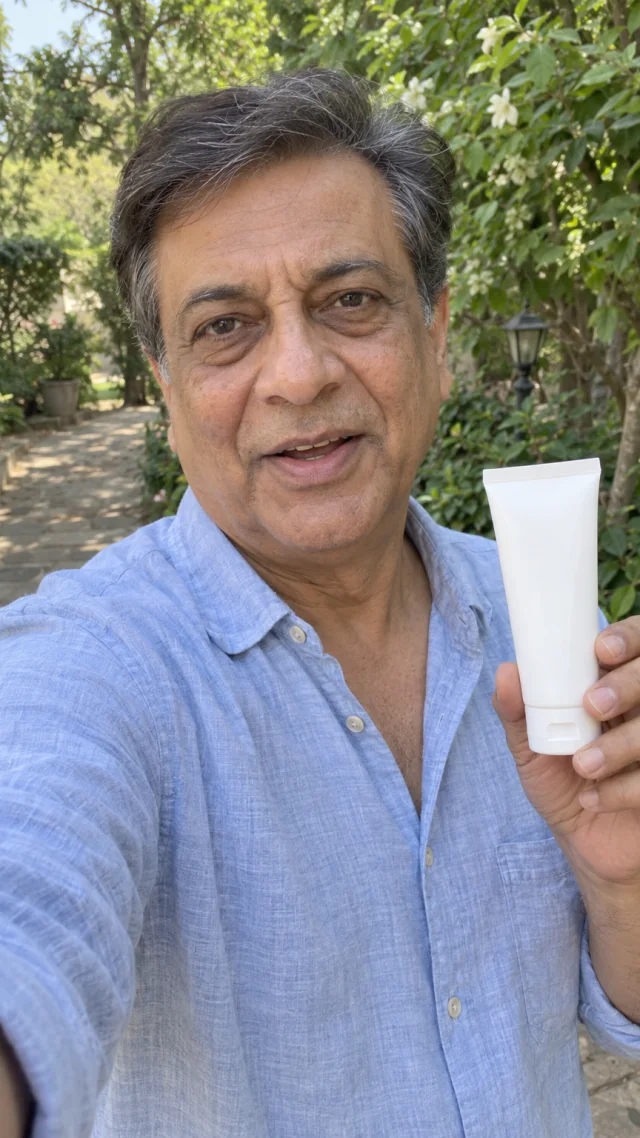 an adult South Asian man in his late fifties in a outdoor setting speaks to camera while holding a plain plain sunscreen tube.