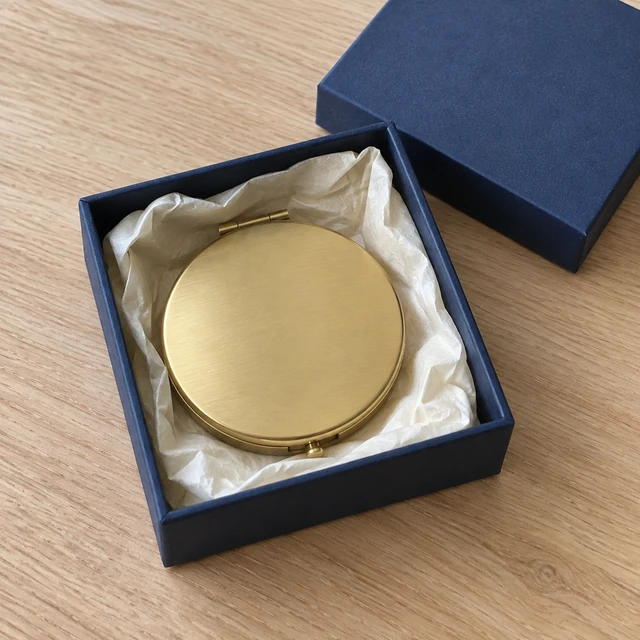 Plain round brass compact mirror nestled in a navy box with cream tissue.