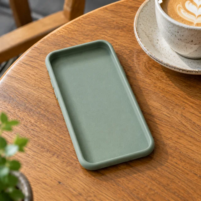 Plain sage silicone phone case lying open on a cafe table beside a coffee cup.