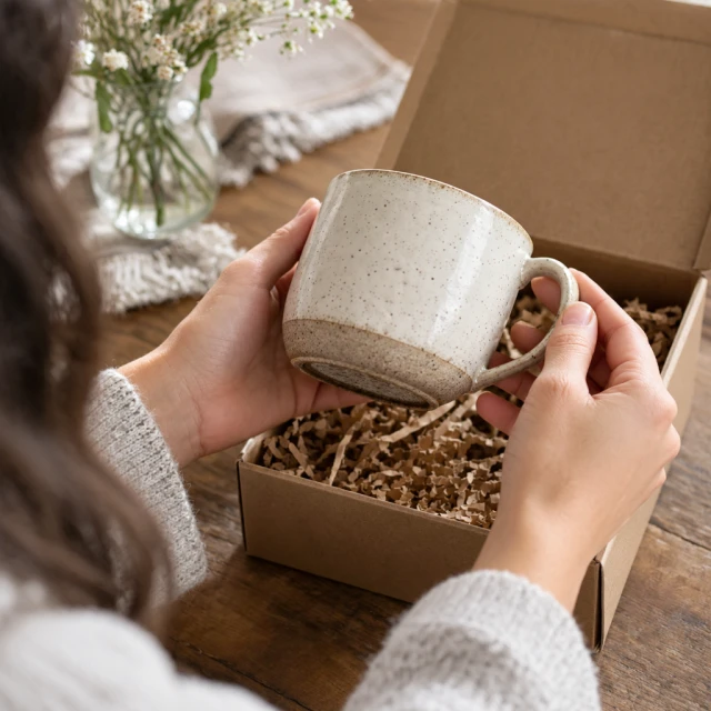 Hands lifting a ceramic mug set out of a gift box with paper filling.