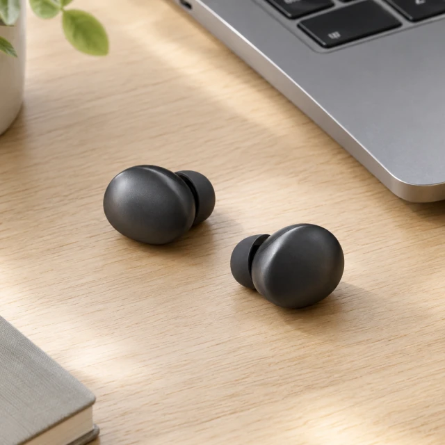 Two rounded black stemless earbuds on a pale oak desk beside a laptop.